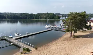 Wdzydze Kiszewskie plaża z pomostami i żaglówkami. Piasek, woda i zielone drzewa tworzą idylliczny krajobraz.