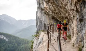 Dwóch rowerzystów pokonuje malownicze trasy rowerowe w Austrii, jadąc wąską ścieżką przy skalnej ścianie z widokiem na góry.