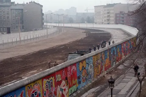 Kolorowy Mur Berliński, świadectwo czasów, kiedy powstał mur berliński, z ludźmi spacerującymi po jego szczycie i budynkami w tle.