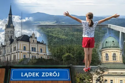 Dziewczynka z rozłożonymi rękami podziwia panoramę Lądka Zdroju z pięknymi budynkami i górami w tle.