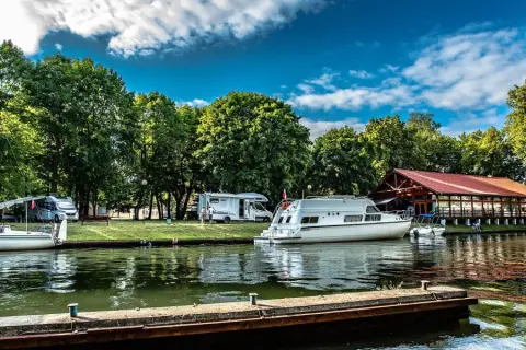 Malownicze pojezierza w Polsce: kampery i łodzie cumują przy brzegu, w tle drewniana restauracja i bujna zieleń.