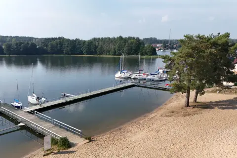Wdzydze Kiszewskie plaża z pomostami i żaglówkami. Piasek, woda i zielone drzewa tworzą idylliczny krajobraz.