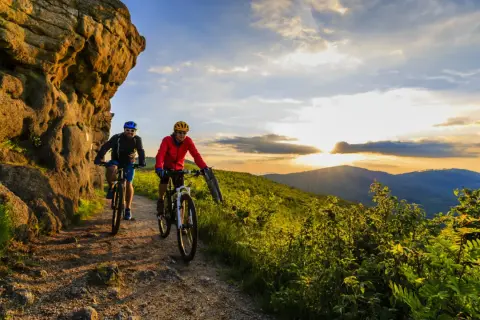 Dwóch rowerzystów przemierza malowniczą ścieżkę, odkrywając najładniejsze trasy rowerowe na Mazowszu. Zachód słońca maluje niebo nad pagórkami.