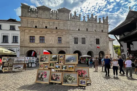 Rynek w Kazimierzu Górnym tętni życiem. Wystawa obrazów z kwiatami i pejzażami przed zabytkową kamienicą.