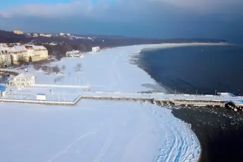 Zimowe atrakcje pomorskie: molo w Sopocie otoczone lodem i śniegiem, z widokiem na zamarznięte morze i nadmorskie budynki.