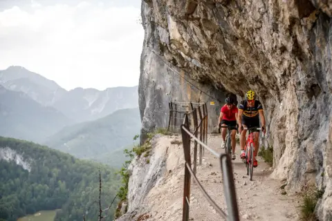 Dwóch rowerzystów pokonuje malownicze trasy rowerowe w Austrii, jadąc wąską ścieżką przy skalnej ścianie z widokiem na góry.