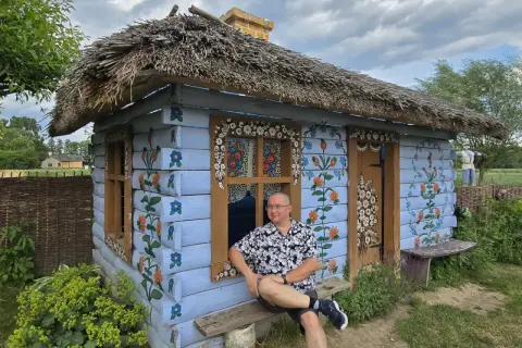 Mężczyzna siedzi na ławce przed malowaną chatką w stylu wsi Zalipie. Chata ozdobiona jest kwiatowymi wzorami.
