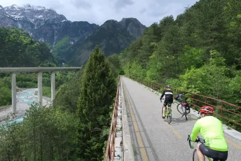 Rowerzyści przemierzają malowniczą trasę **Alpe Adria** w otoczeniu gór i lasów. Widoczny most nad rzeką dodaje krajobrazowi majestatu.