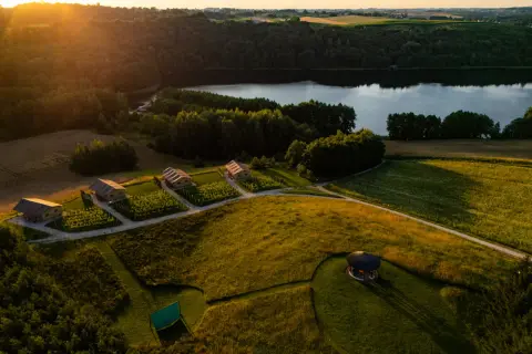 Idealny weekend w Polsce: domki glampingowe nad jeziorem, otoczone zielenią i polami. Słońce zachodzi, rzucając złote światło na krajobraz.