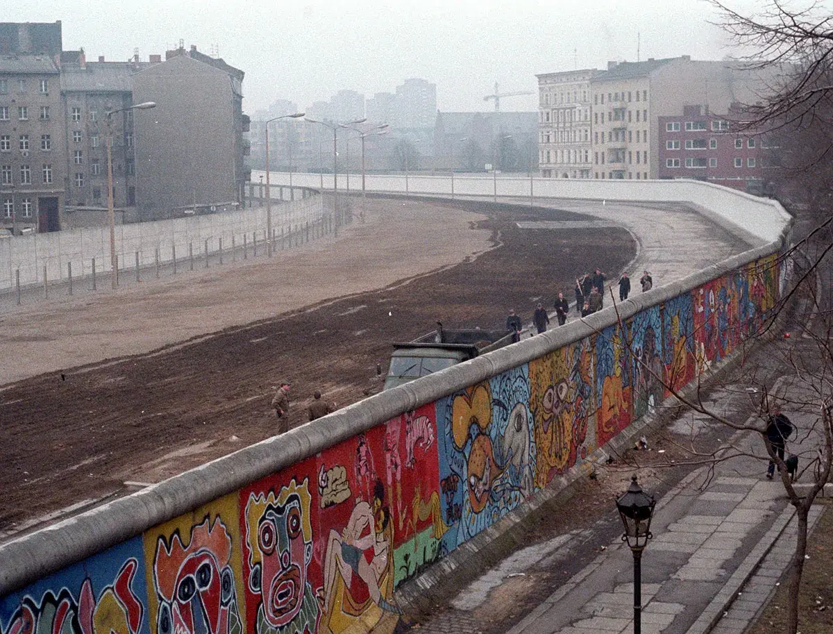 Kolorowy Mur Berliński, świadectwo czasów, kiedy powstał mur berliński, z ludźmi spacerującymi po jego szczycie i budynkami w tle.