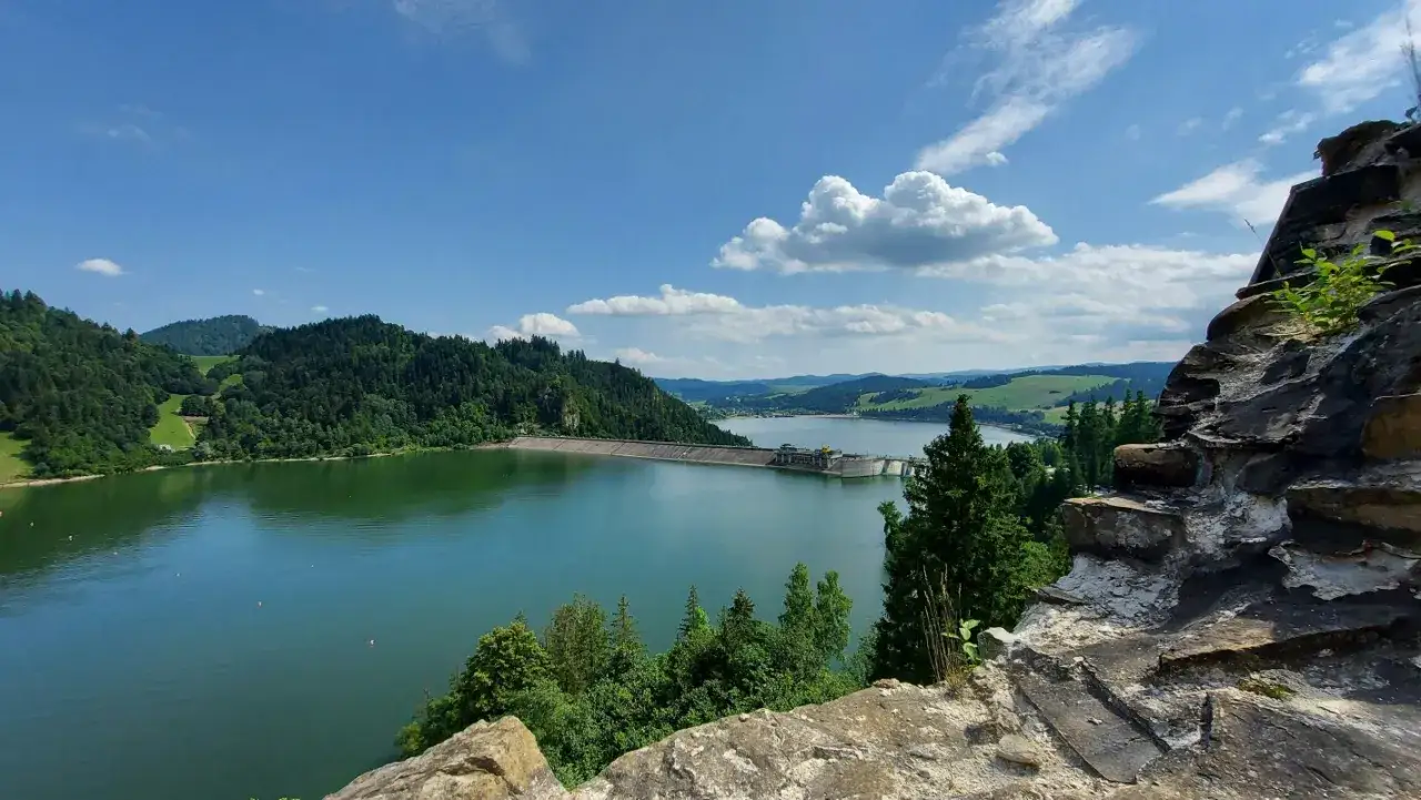 Piękne jezioro w górach, otoczone zielonymi lasami, z tamą w tle. Błękitne niebo z białymi chmurami.