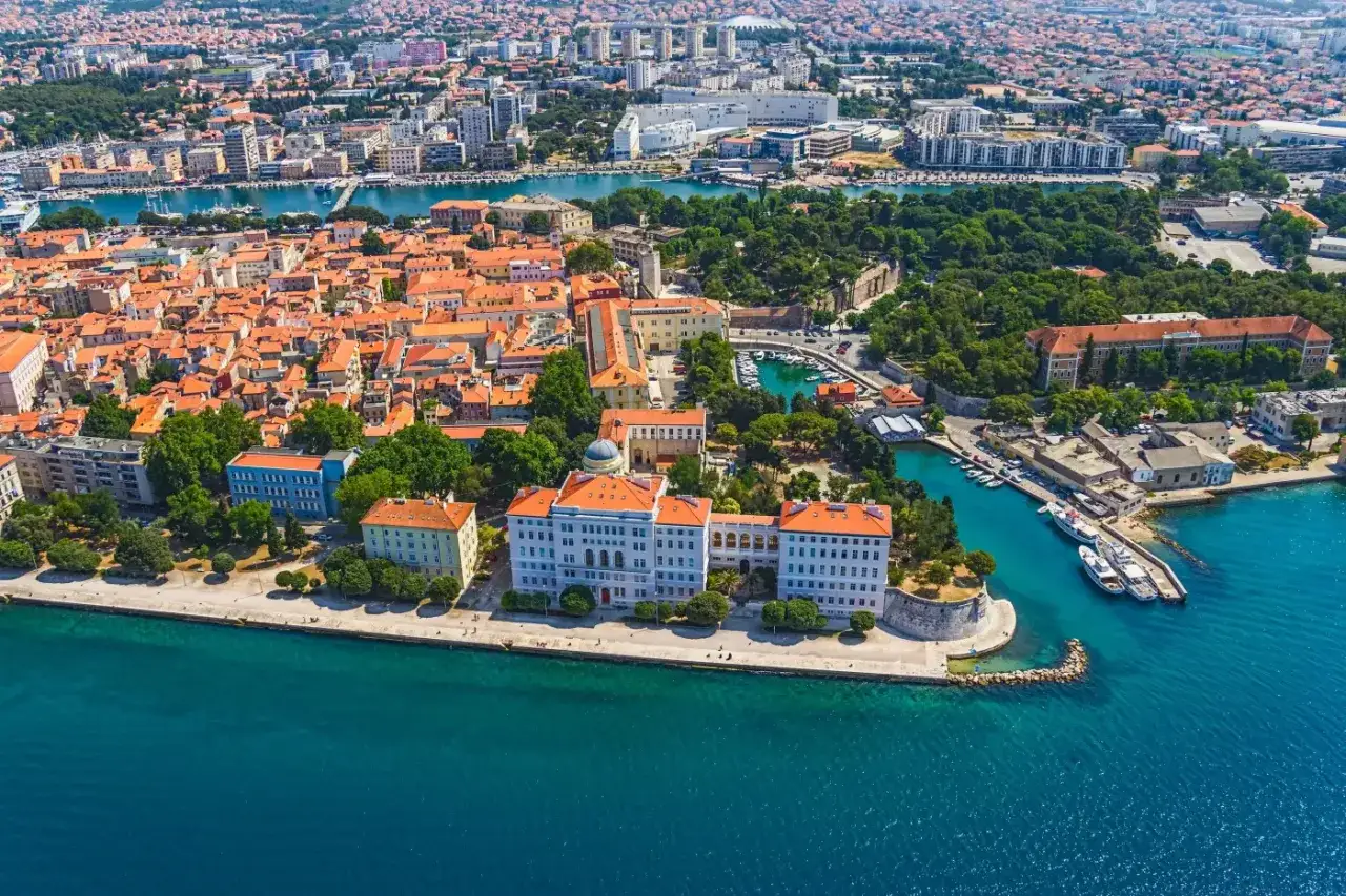 Widok z lotu ptaka na Zadar, z jego pomarańczowymi dachami i turkusowym morzem. Miasto pięknie prezentuje się na tle błękitnego nieba, oferując widok, który może konkurować ze Splitem.