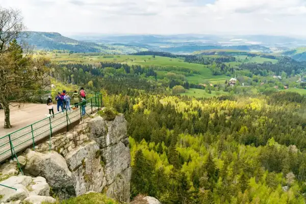 Widok na malownicze szczyty Kotliny Kłodzkiej z platformy widokowej. Grupa turystów podziwia rozległą panoramę lasów i zielonych wzgórz.